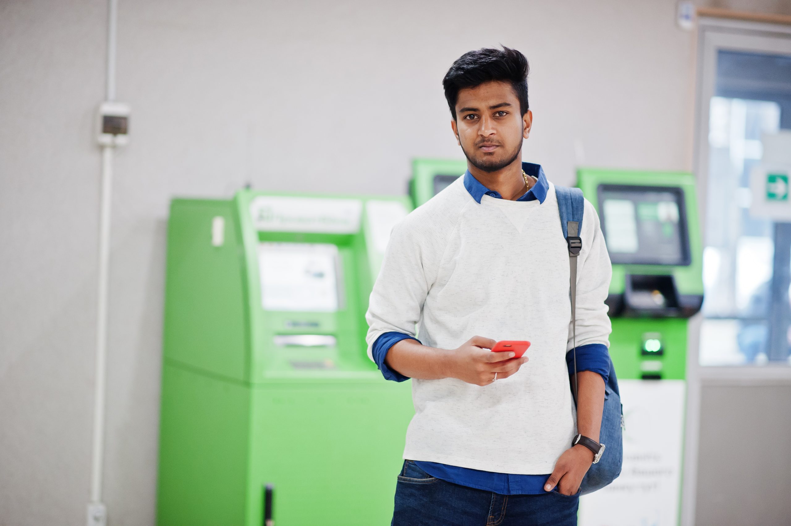 Young stylish asian man with mobile phone and backpack against row of green ATM.